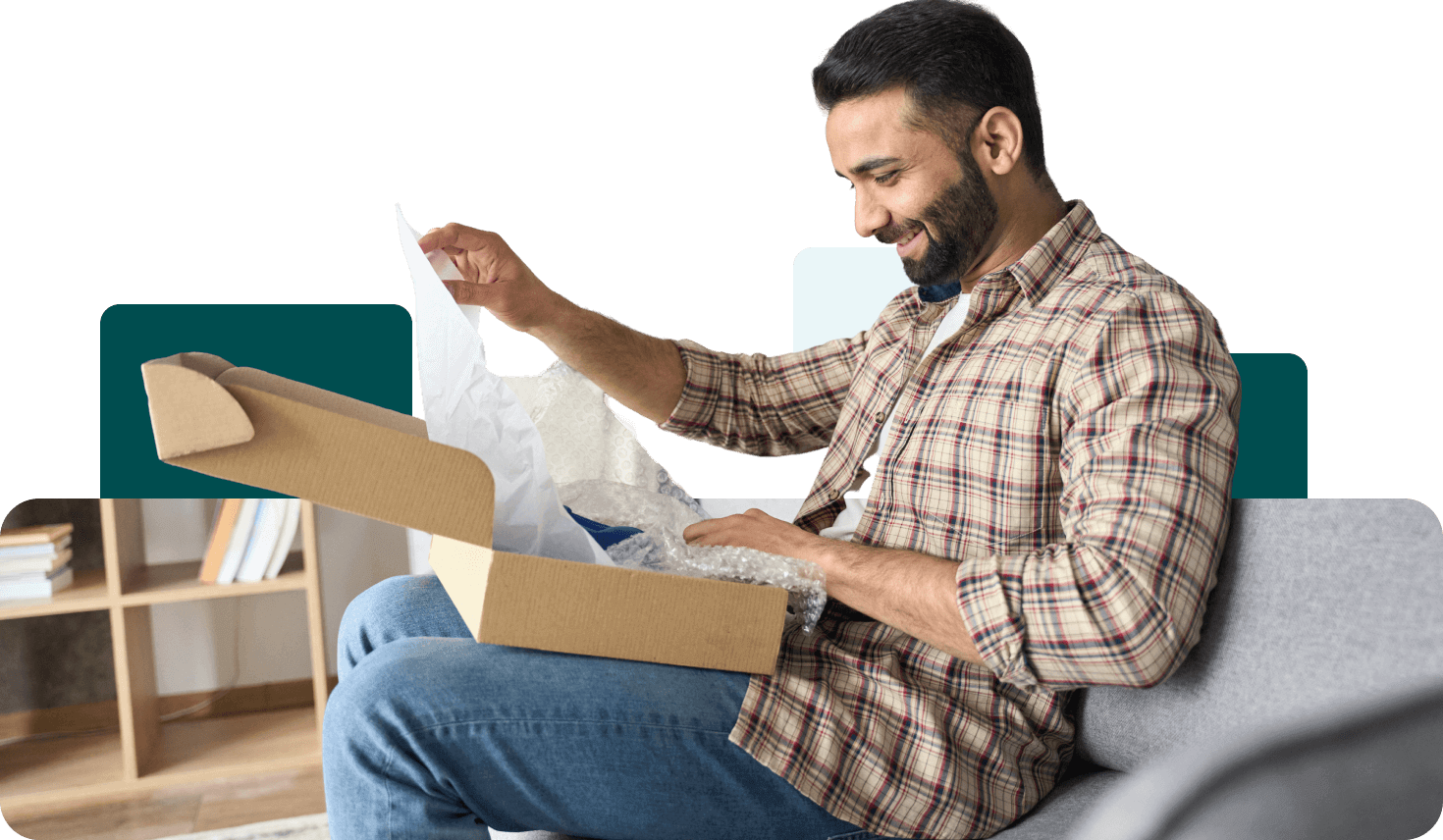 A man sits on a couch, wearing a plaid shirt, and smiles while opening a cardboard box with bubble wrap inside.